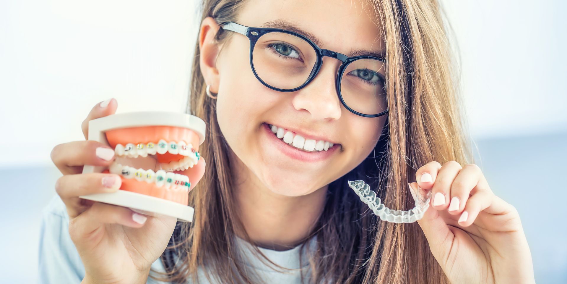 imagem de jovem menina usando óculos e a sorrir com representação de aparelho dentário fixo numa mão e a segurar alinhador de invisalign noutra mão.