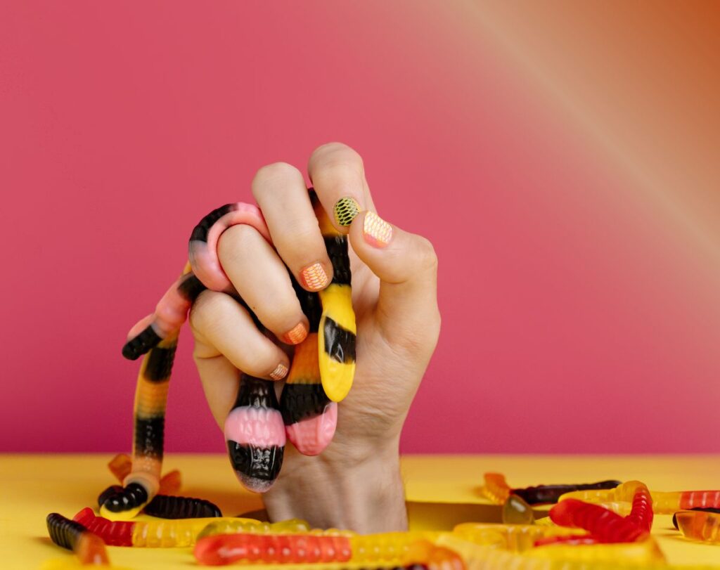 imagem de mão com unhas pintadas de diferentes cores a agarrar minhocas de goma